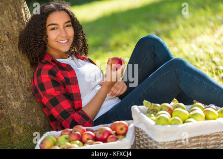 Beautiful happy mixed race African American girl teenager female young woman in an orchard eating an organic red apple, smiling with perfect teeth lea Stock Photo