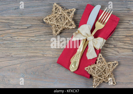 Christmas table place setting. Gold cutlery, red napkin, and gold stars with festive ribbon on rustic wood. Stock Photo