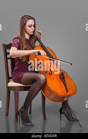 Beautiful young woman sitting on the chair and playing cello. Isolated on grey background Stock Photo