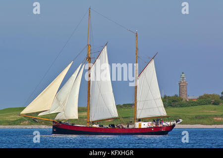 Historic two-masted sailing ship at sea with green shore and grey stone lighthouse in the background Stock Photo