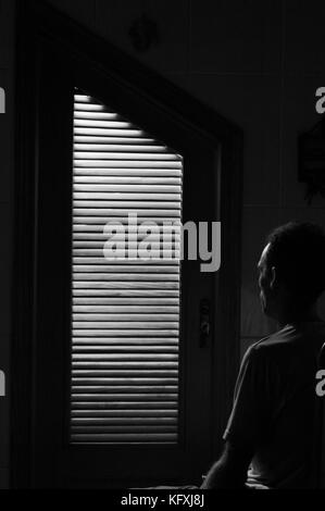 Door of cupboard with flushed light within illuminating a seated man looking at the door. Black and white image.