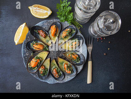 Mussels baked with butter and parsley in shell mussels. Lemon, parsley and spices around plate on dark background. Top view. Healthy eating concept Stock Photo