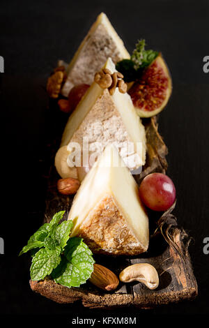 Pieces of moldy cheese with grape, mint, figs, almonds, cashews on bark on black background. Food styling composition with dried cheese. Stock Photo