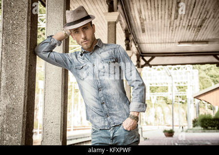 Portrait of blond haired handsome young man with fedora hat. An old train station is behind him in Italian town Stock Photo