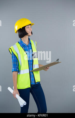 Smiling female architect holding clipboard and blueprint against grey background Stock Photo