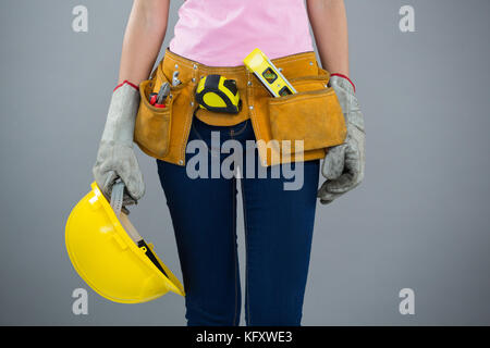 Mid section of woman with tool belt and holding hard hat against grey background Stock Photo