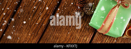 Snow falling against pine cones and wrapped gift box on wooden table Stock Photo
