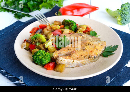 Steaks of fried spicy fish, served with a warm vegetable salad on a ceramic plate on a white wooden background. Stock Photo