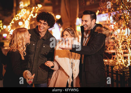 Cheerful four friends having fun in city street in Christmas night, talking something funny. Stock Photo