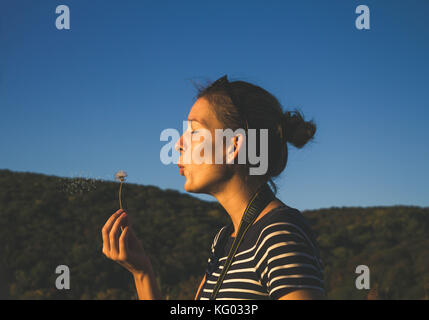 A standing young girl blowing dandelion flower, green mountain forests in background Stock Photo