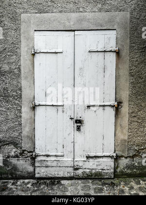 Vintage painted white wood double door with stucco frame, strap hinges, and padlock with a rustic and worn look and cobblestone road in foreground Stock Photo