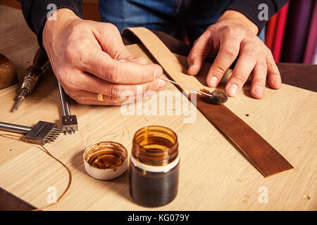 A close-up of a furrier paints a piece of genuine leather in brown for making a belt on the desktop, on which stands a can of paint Stock Photo