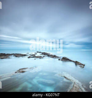 Dark rocks in a blue ocean under cloudy sky in a bad weather. Long exposure photography Stock Photo