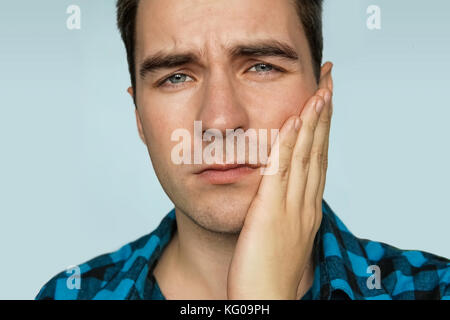 A young guy with a sad emotion on her face holding his hand over the patient's jaw on a blue background. Portrait of a man in painful expression with  Stock Photo