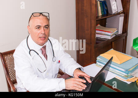 Doctor in his studio at desk, on pc,  with glasses. Use new technologies. In his professional studio, he is sitting at antique desk, and a green lamp  Stock Photo