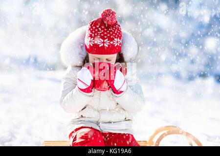 Child drinking hot chocolate with marshmallows in snowy winter park. Kid with cup of warm cocoa drink on Christmas vacation. Little girl playing in sn Stock Photo