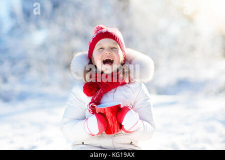 Child drinking hot chocolate with marshmallows in snowy winter park. Kid with cup of warm cocoa drink on Christmas vacation. Little girl playing in sn Stock Photo