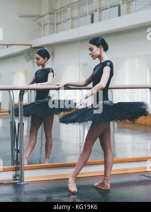 Ballerina in black tutu and pointe stretches on barre in ballet gym. Woman standing near bar and mirror, preparing for perfomance. Stock Photo