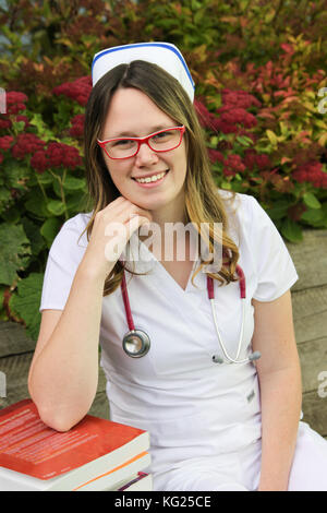 Young smiling female nurse portrait wearing white scrubs, cap and a stethoscope around her neck leaning on a stack of books outdoors during the Fall Stock Photo