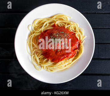Cooked spaghetti pasta dish, hot with steam coming out, with red tomato sauce and oregano sprinkled on top, on a black wooden table background Stock Photo