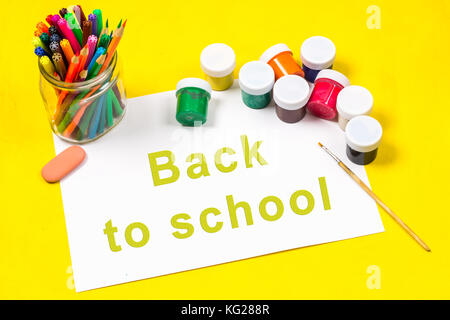 The inscription - back to school, on a sheet of white paper surrounded by colored pencils, markers, paints on a bright yellow background Stock Photo
