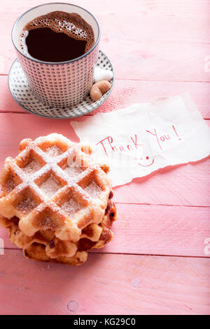 Cute thank you note on a piece of paper with a smiley face on it,  on a pink background, near a hot cup of coffee and fresh waffles, with powdered sug Stock Photo