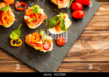 Canapes appetizer with prosciutto ham, pepper slices, tomatoes and green spinach and rucola leaves, healthy crostini or bruschettas italian appetizer  Stock Photo