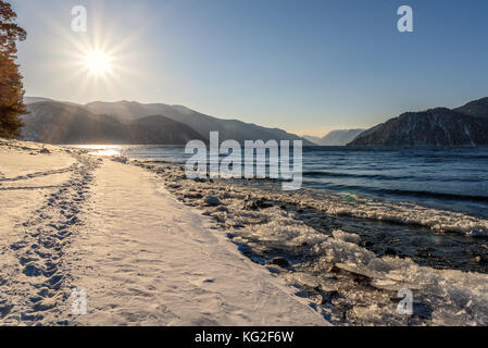 Beautiful winter landscape with a lake, mountains, sun and footprints on the snow along the shore against a blue sky Stock Photo