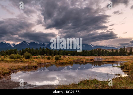Scenic view with mountains, forest, dirt road through ford, storm clouds and reflections on a background of cloudy sky at sunset