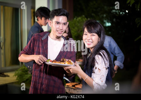 Asian young couple enjoying a romantic dinner and  group of friends having outdoor garden barbecue laughing with alcoholic beer drinks on night Stock Photo