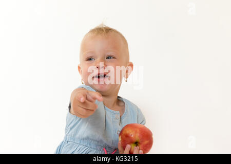 Studio shot of sitting cool toddler girl holding red apple and pointing her finger at the camera Stock Photo