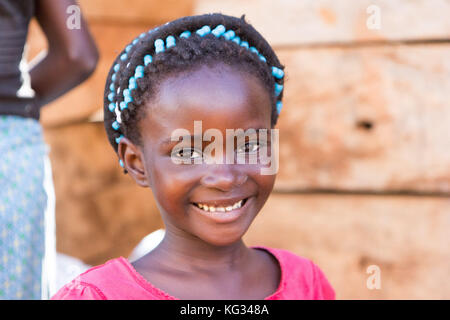 A smiling girl with colorful beads on her hair braids, she is posing in front of a wooden shack. Stock Photo