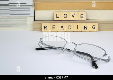 The words love reading written in letter tiles resting against a pile of books and novels on a white surface with some silver glasses (spectacles) Stock Photo