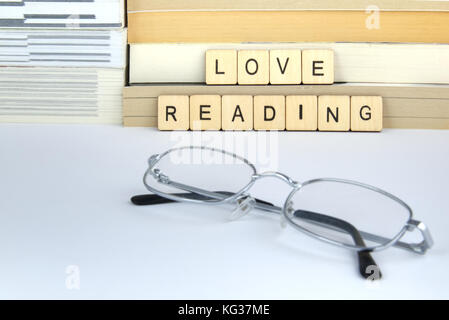 The words love reading written in letter tiles resting against a pile of books and novels on a white surface with some silver glasses (spectacles) Stock Photo