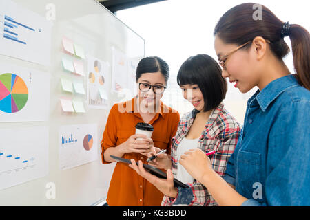 cheerful pretty business women team partner standing in front of whiteboard and looking at mobile digital tablet discussing design case plan. Stock Photo