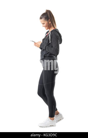 Side view of young happy fit runner woman using mobile phone and smiling. Full body length portrait isolated on white background. Stock Photo