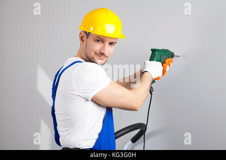 Young man in yellow protective helmet with electric drill making hole in wall Stock Photo