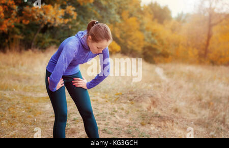 Exhausted caucasian jogger girl in blue shirt and black sports leggins resting after jogging on the colorful autumnal forest hill. Stock Photo