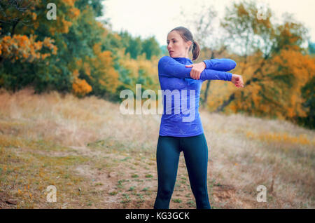 Slim caucasian jogger girl in blue shirt and black sports leggins performs warming-up with hands  before jogging on the yellow autumnal forest hill. Stock Photo