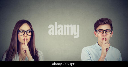Secretive people. Young woman and man looking at each other saying hush be quiet with finger on lips gesture isolated on gray wall background. Stock Photo