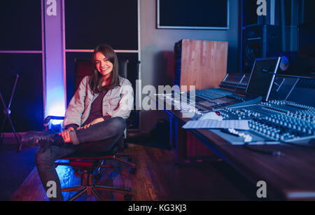 Happy young woman sitting in sound recording studio. Female music composer sitting by sound mixing console. Stock Photo