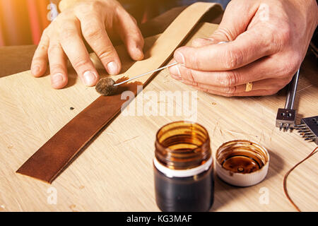 A close-up of a furrier paints a piece of genuine leather in brown for making a belt on the desktop, on which stands a can of paint Stock Photo