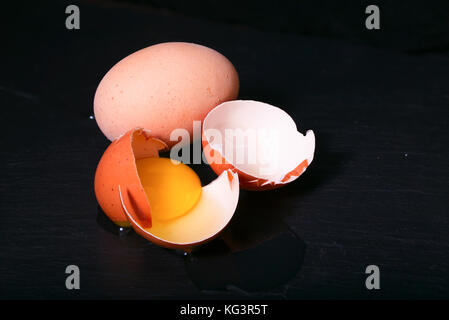 The broken egg against a dark background. Two eggs, a single whole, the second broken have also partially flowed out on a black table Stock Photo