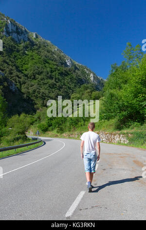 The person on the highway in mountains. The young man goes along the highway, between the mountain tops covered with green trees. Summer sunny day, th Stock Photo