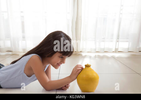 Little girl lying on floor and putting coin in clay piggy bank Stock Photo