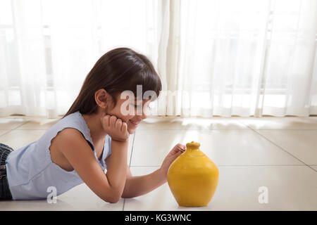 Little girl lying on floor and putting coin in clay piggy bank Stock Photo