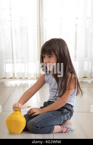 Little girl sitting on floor and putting bank note in clay piggy bank Stock Photo