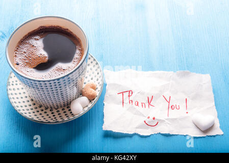 Paper note with a thank you message and a smiley face, a cup of hot aromatic coffee with pieces of heart shaped sugar, on a blue wooden table, on a su Stock Photo