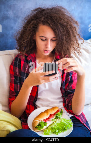 Curly hair teen girl using mibile phone and eating sandwich on sofa Stock Photo