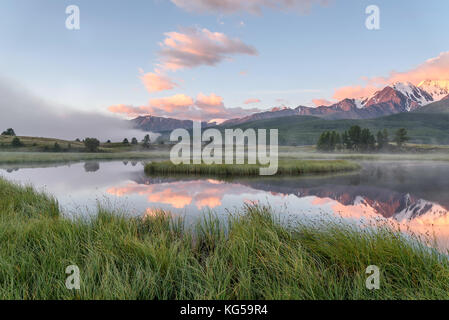 Picturesque pink dawn with a beautiful mist over the lake, mountains covered with snow, green trees, grass, blue sky, clouds and their reflections Stock Photo